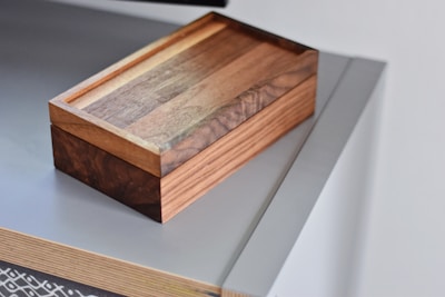 A wooden box with a polished surface rests on a modern, sleek tabletop. The box has a rich brown color with visible wood grain, highlighting its craftsmanship. The tabletop appears to be a minimalist design with a light grey color and a smooth finish.