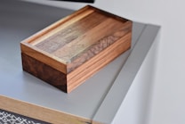 A wooden box with a polished surface rests on a modern, sleek tabletop. The box has a rich brown color with visible wood grain, highlighting its craftsmanship. The tabletop appears to be a minimalist design with a light grey color and a smooth finish.