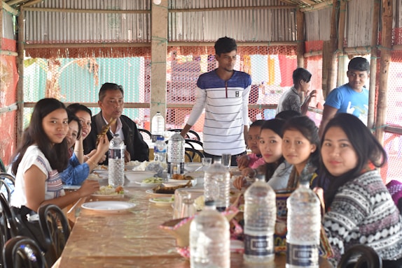 A group of people are seated around a table, sharing a meal in a rustic setting. The table is covered with a light brown cloth and various dishes are placed on it. Multiple clear water bottles are visible on the table. The setting appears informal and friendly, with a structure made of wooden beams and metal sheeting as the backdrop.