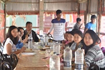 A group of people are seated around a table, sharing a meal in a rustic setting. The table is covered with a light brown cloth and various dishes are placed on it. Multiple clear water bottles are visible on the table. The setting appears informal and friendly, with a structure made of wooden beams and metal sheeting as the backdrop.