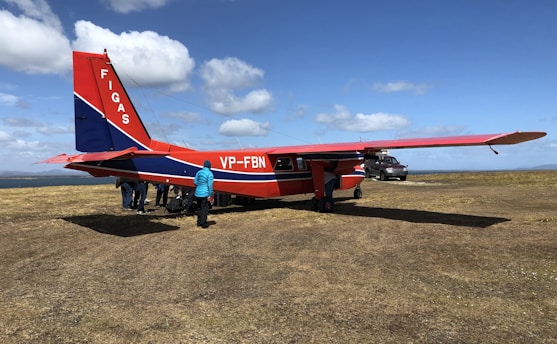 A small red and blue airplane is stationary on a grass-covered airfield. A group of people is gathered around the aircraft, with one person wearing a bright blue jacket standing prominently near the plane. The sky above is partly cloudy, creating a bright and clear weather ambiance. A pickup truck is parked in the background, adding to the rural and open setting.