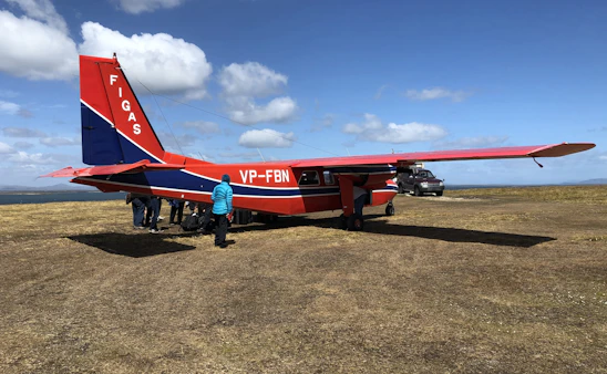A welcoming group of pilots and instructors standing beside small aircraft on a sunny airfield.