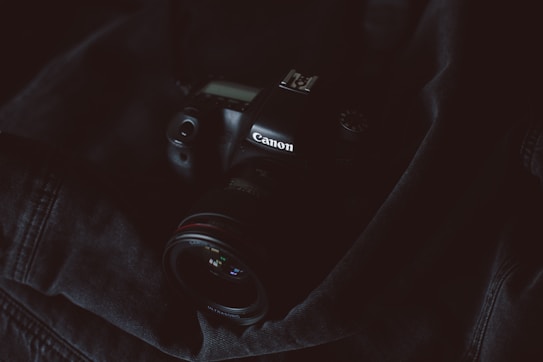 A Canon DSLR camera rests on a dark, textured fabric. The camera, which is partially illuminated, contrasts with the surrounding shadows. The lens is prominent, and some of the camera's dials and buttons are visible, adding to the sense of detail and complexity in the equipment.
