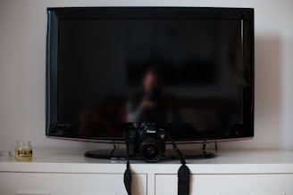 A flat-screen TV is positioned on top of a white cabinet. In front of the TV, there's a digital camera resting on its stand. To the left of the TV, a small candle jar can be seen.