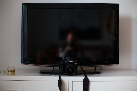 A flat-screen TV is positioned on top of a white cabinet. In front of the TV, there's a digital camera resting on its stand. To the left of the TV, a small candle jar can be seen.