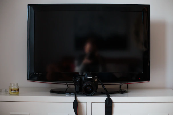 A flat-screen TV is positioned on top of a white cabinet. In front of the TV, there's a digital camera resting on its stand. To the left of the TV, a small candle jar can be seen.