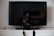 A flat-screen TV is positioned on top of a white cabinet. In front of the TV, there's a digital camera resting on its stand. To the left of the TV, a small candle jar can be seen.