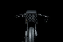 A dark and minimalistic image featuring the front view of a motorcycle, highlighting the intricate honeycomb pattern on the front cover. The handlebars and the textured tire are also visible, set against a black background.