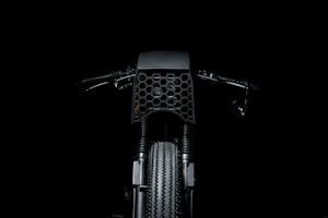 A dark and minimalistic image featuring the front view of a motorcycle, highlighting the intricate honeycomb pattern on the front cover. The handlebars and the textured tire are also visible, set against a black background.