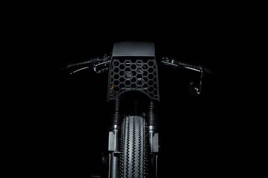 A dark and minimalistic image featuring the front view of a motorcycle, highlighting the intricate honeycomb pattern on the front cover. The handlebars and the textured tire are also visible, set against a black background.
