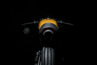 A motorcycle is illuminated against a dark background. The focus is on the rear view showing the tire and fender, with handlebars visible to the sides. The motorcycle has a sleek design with a mix of modern and classic elements. The dominant color is a bright yellow contrasted with black.