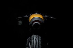 A motorcycle is illuminated against a dark background. The focus is on the rear view showing the tire and fender, with handlebars visible to the sides. The motorcycle has a sleek design with a mix of modern and classic elements. The dominant color is a bright yellow contrasted with black.