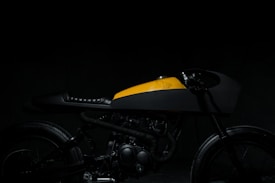 A sleek and stylized motorcycle is partially visible, with a yellow and black color scheme. The focus is on the body and fuel tank, displaying a minimalist design with a vintage feel. The lighting is dim, creating dramatic shadows and highlighting the contours of the bike.