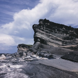 A dramatic rocky cliff with weathered layers stands against a partly cloudy sky. The rugged terrain is composed of dark and light striations, contrasting with the sky's soft blues and whites. The scene conveys a sense of grandeur and natural beauty.