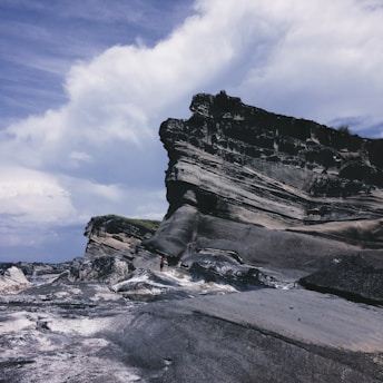 A dramatic rocky cliff with weathered layers stands against a partly cloudy sky. The rugged terrain is composed of dark and light striations, contrasting with the sky's soft blues and whites. The scene conveys a sense of grandeur and natural beauty.