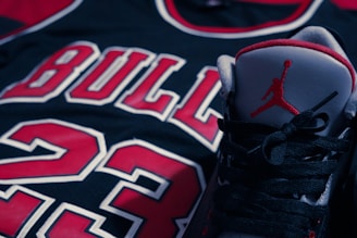 A close-up shot of a black basketball jersey with the number 23 and the word 'BULLS' in red and white lettering. An athletic shoe featuring a red silhouette of a basketball player is positioned prominently in the foreground.