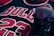 A close-up shot of a black basketball jersey with the number 23 and the word 'BULLS' in red and white lettering. An athletic shoe featuring a red silhouette of a basketball player is positioned prominently in the foreground.
