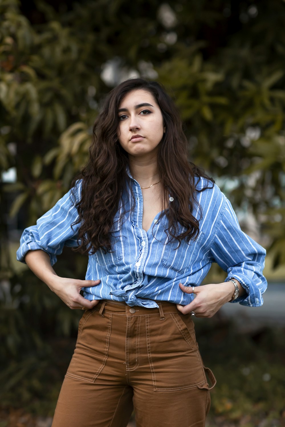 woman wearing blue and white striped button-up long-sleeved shirt and brown pants standing near tree during daytime