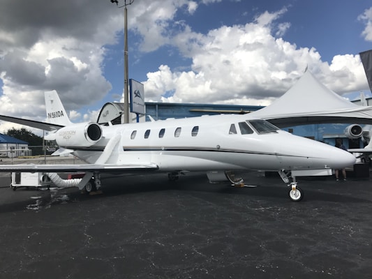 A sleek white private jet is parked on the tarmac under partly cloudy skies. The aircraft features two engines and several small windows along the fuselage. A white tent and a blue building are visible in the background, along with a flagpole.