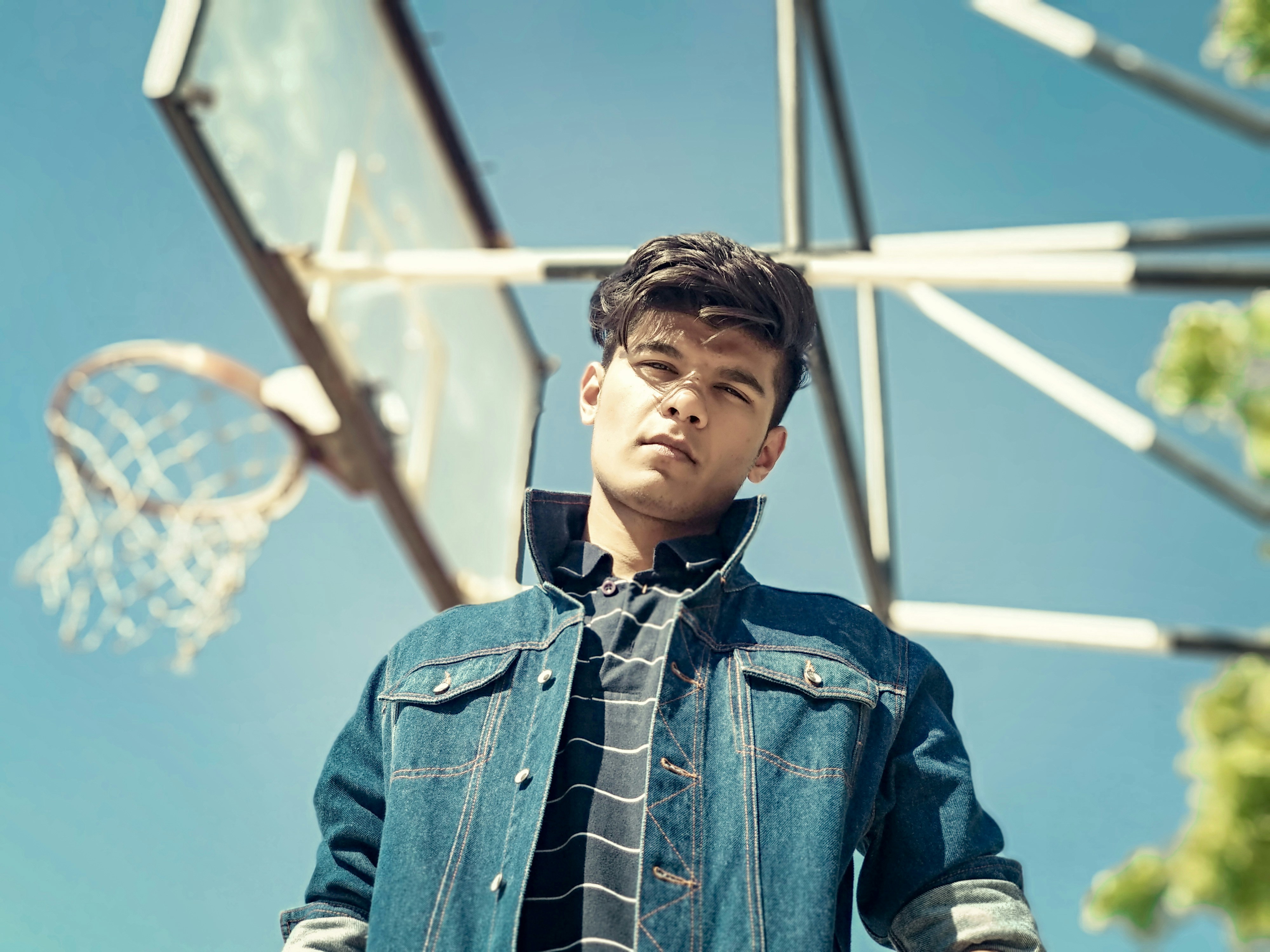 low angle photography of standing man wearing black-and-white striped shirt and blue denim button-up jacket beside white basketball system during daytime