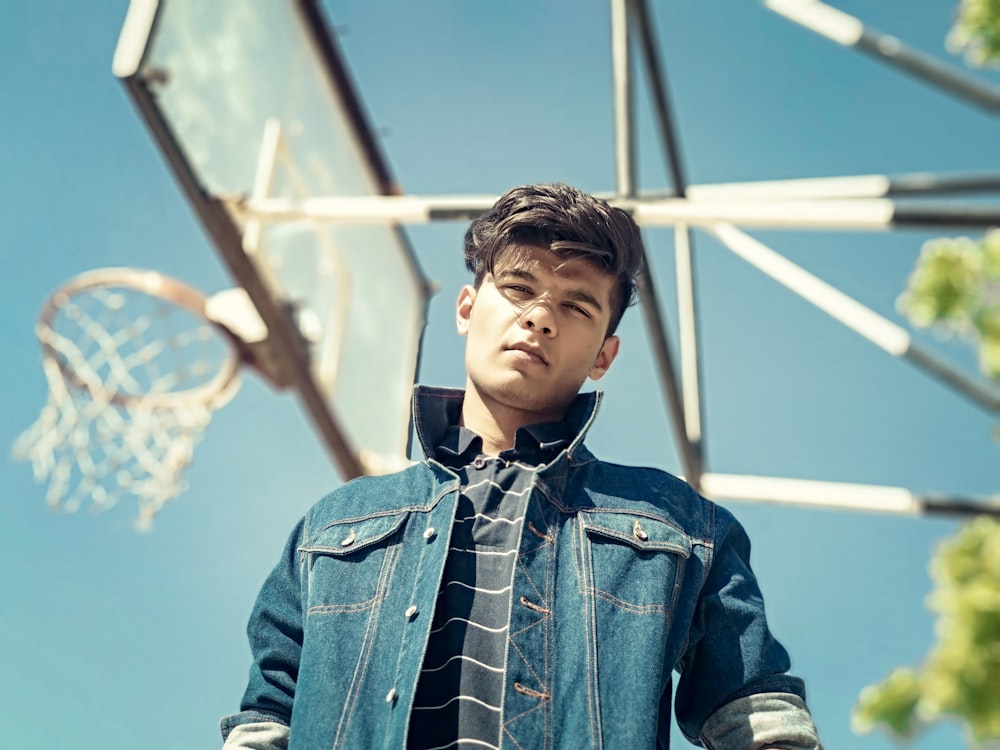 low angle photography of standing man wearing black-and-white striped shirt and blue denim button-up jacket beside white basketball system during daytime