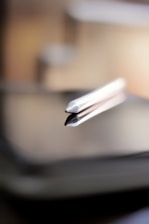 A close-up view of a stylus pen resting on the surface of a digital tablet. The focus is sharp on the tip of the stylus, while the background and parts of the tablet are softly blurred, creating a depth-of-field effect. The lighting is warm and emphasizes the sleek, metallic design of the stylus.