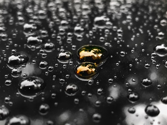 Raindrops are scattered across a reflective surface, each drop varying in size. The surface is dark, and the drops create a pattern that appears almost metallic. A few of the drops reflect a warm, golden hue, possibly from surrounding light sources.