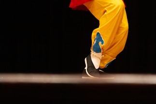 A close-up of a person's legs dressed in vibrant yellow pants with blue decorative patterns. The shoes are black with white soles, and the person seems to be performing a step or dance move against a dark background.