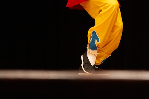 A close-up of a person's legs dressed in vibrant yellow pants with blue decorative patterns. The shoes are black with white soles, and the person seems to be performing a step or dance move against a dark background.