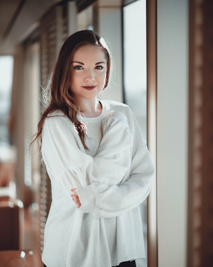 A person with long brown hair and a white sweater stands by a window, capturing natural light. The setting appears indoors with a soft, neutral background, giving a cozy and serene atmosphere.
