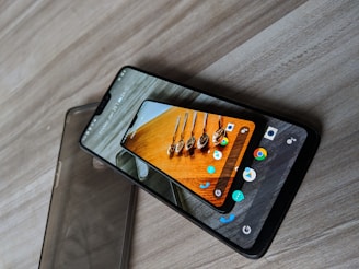 A smartphone with a protective case partially slipped off is placed on a wooden surface. The phone's screen displays a colorful image of spoons containing various spices. Multiple app icons are visible around the image, and at the top of the screen, the date and weather information can be seen.