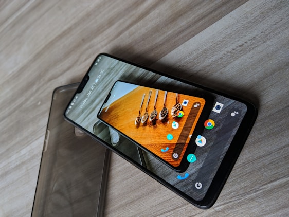 A smartphone with a protective case partially slipped off is placed on a wooden surface. The phone's screen displays a colorful image of spoons containing various spices. Multiple app icons are visible around the image, and at the top of the screen, the date and weather information can be seen.