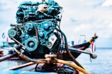 A close-up of a boat engine with various mechanical components like belts and gears, set against a backdrop of a boat on water, with some out-of-focus elements suggesting a maritime setting.