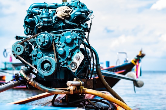A close-up of a boat engine with various mechanical components like belts and gears, set against a backdrop of a boat on water, with some out-of-focus elements suggesting a maritime setting.