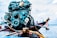 A close-up of a boat engine with various mechanical components like belts and gears, set against a backdrop of a boat on water, with some out-of-focus elements suggesting a maritime setting.