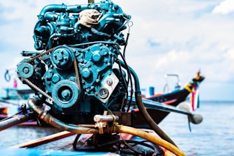 A close-up of a boat engine with various mechanical components like belts and gears, set against a backdrop of a boat on water, with some out-of-focus elements suggesting a maritime setting.