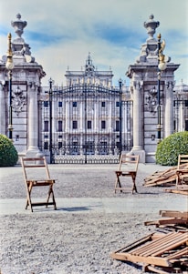 A grand, ornate gate with iron bars is flanked by two decorative pillars topped with elaborate urns. Behind the gate, a majestic building with classical architecture is visible. In the foreground, there are a few wooden chairs scattered on a gravel ground, and some bushes can be seen on either side of the entrance.