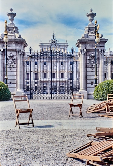 A grand, ornate gate with iron bars is flanked by two decorative pillars topped with elaborate urns. Behind the gate, a majestic building with classical architecture is visible. In the foreground, there are a few wooden chairs scattered on a gravel ground, and some bushes can be seen on either side of the entrance.