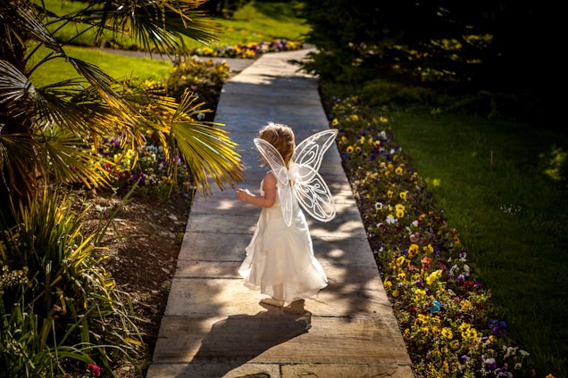 A young child wearing fairy wings and a white dress walks down a garden path bordered by colorful flowers. Sunlight filters through the trees, casting playful shadows on the stone walkway.