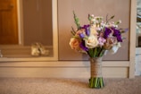 A beautifully arranged bouquet of flowers sits on a carpeted floor in front of a wooden cabinet. The bouquet features a mix of cream, pink, and purple flowers with greenery, all tied together with a burlap wrap. A pair of blurred shoes is visible in the background, suggesting a bridal or formal setting.