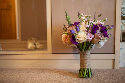 A beautifully arranged bouquet of flowers sits on a carpeted floor in front of a wooden cabinet. The bouquet features a mix of cream, pink, and purple flowers with greenery, all tied together with a burlap wrap. A pair of blurred shoes is visible in the background, suggesting a bridal or formal setting.
