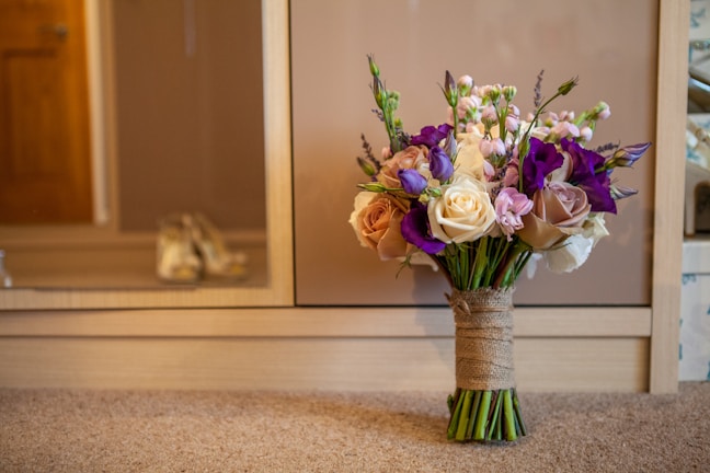 A beautifully arranged bouquet of flowers sits on a carpeted floor in front of a wooden cabinet. The bouquet features a mix of cream, pink, and purple flowers with greenery, all tied together with a burlap wrap. A pair of blurred shoes is visible in the background, suggesting a bridal or formal setting.