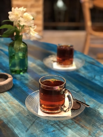 A cozy setting features a blue wooden table with two glass cups of tea, each on a clear saucer with a napkin and a cookie beside them. A green bottle vase with white flowers adds a touch of elegance to the scene, while blurred wooden and brick textures in the background create a warm ambiance.