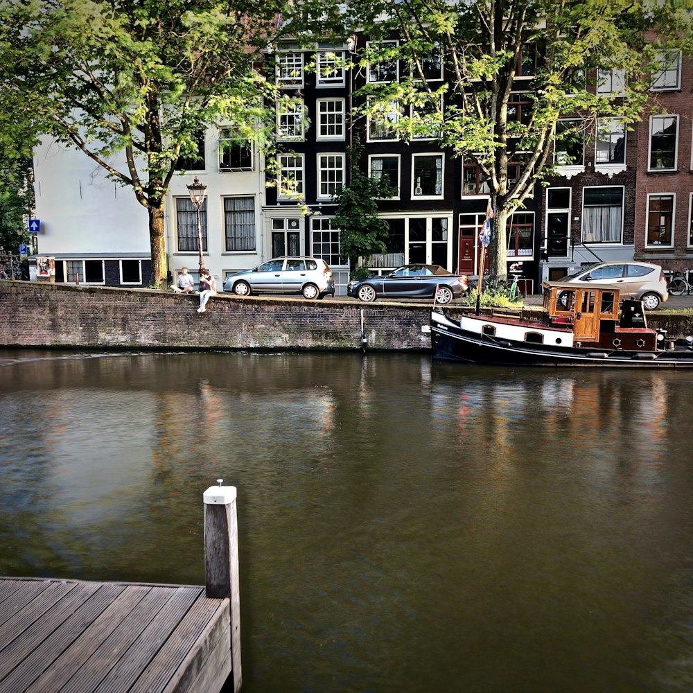 two person sitting on ledge at river with brown and black boat and cars parked beside road during daytime