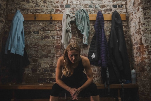 A person sits pensively on a wooden bench in a dimly lit locker room with a brick wall. Several coats and garments hang on hooks above the bench, and a water bottle is placed on the side.