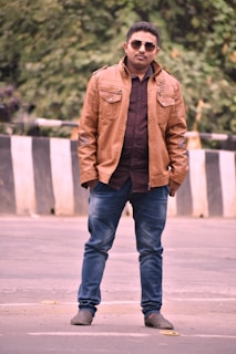 A man stands confidently on a street wearing a brown leather jacket, blue jeans, and sunglasses. The background features greenery and a black-and-white striped barrier.