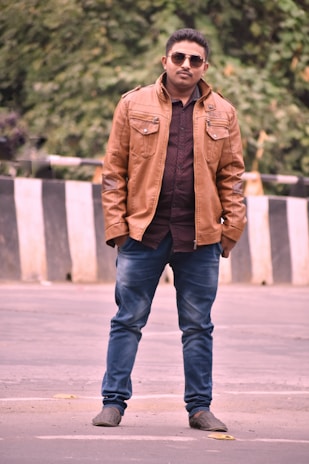 A man stands confidently on a street wearing a brown leather jacket, blue jeans, and sunglasses. The background features greenery and a black-and-white striped barrier.
