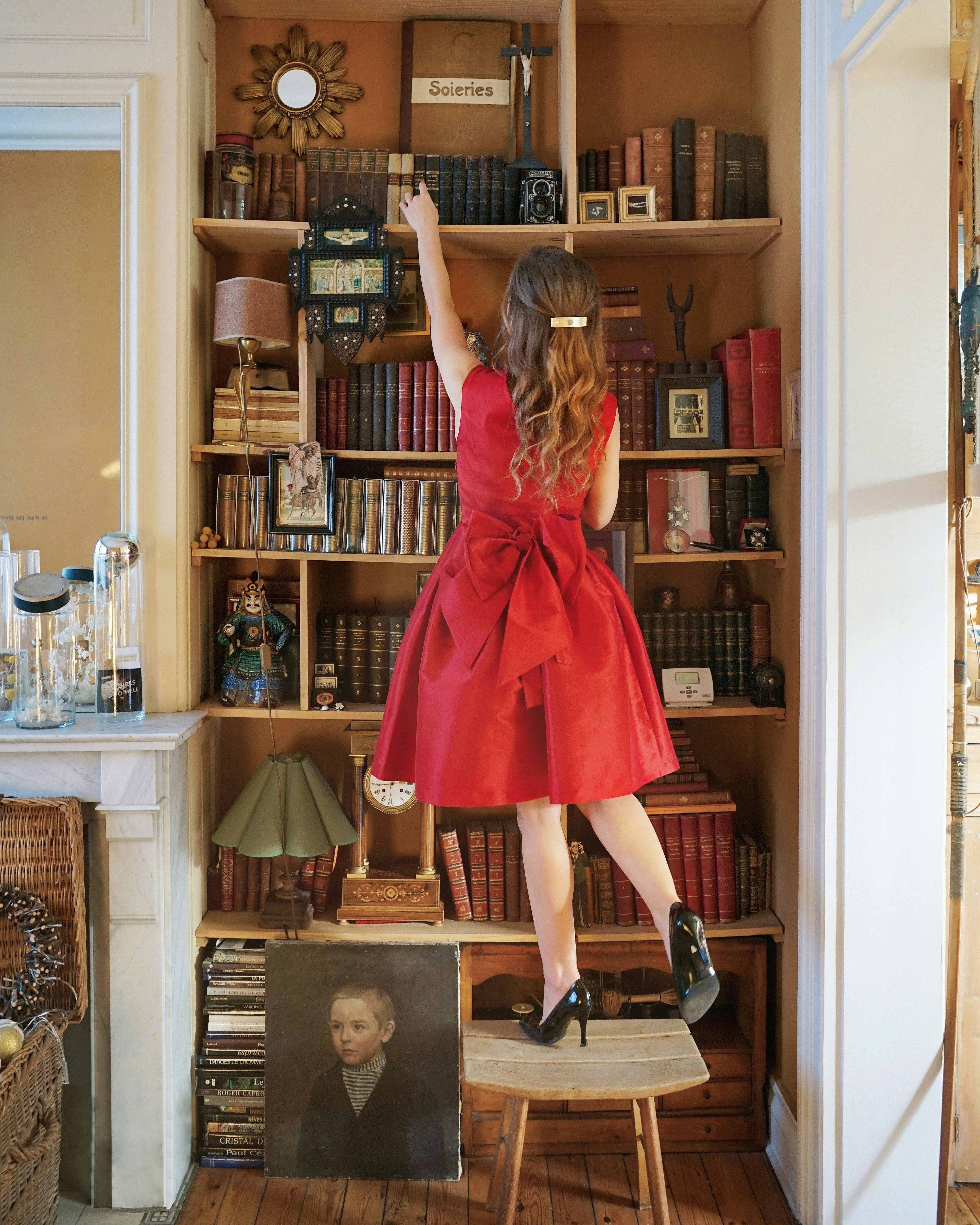 girl in re dress standing on stool while reaching books on shelve