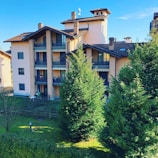 A multi-story residential building with brown roofs and multiple balconies, surrounded by lush green trees and a well-maintained lawn. The sky is clear and blue, contributing to a peaceful atmosphere, and there is a small fenced area in front of the building.