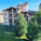 A multi-story residential building with brown roofs and multiple balconies, surrounded by lush green trees and a well-maintained lawn. The sky is clear and blue, contributing to a peaceful atmosphere, and there is a small fenced area in front of the building.